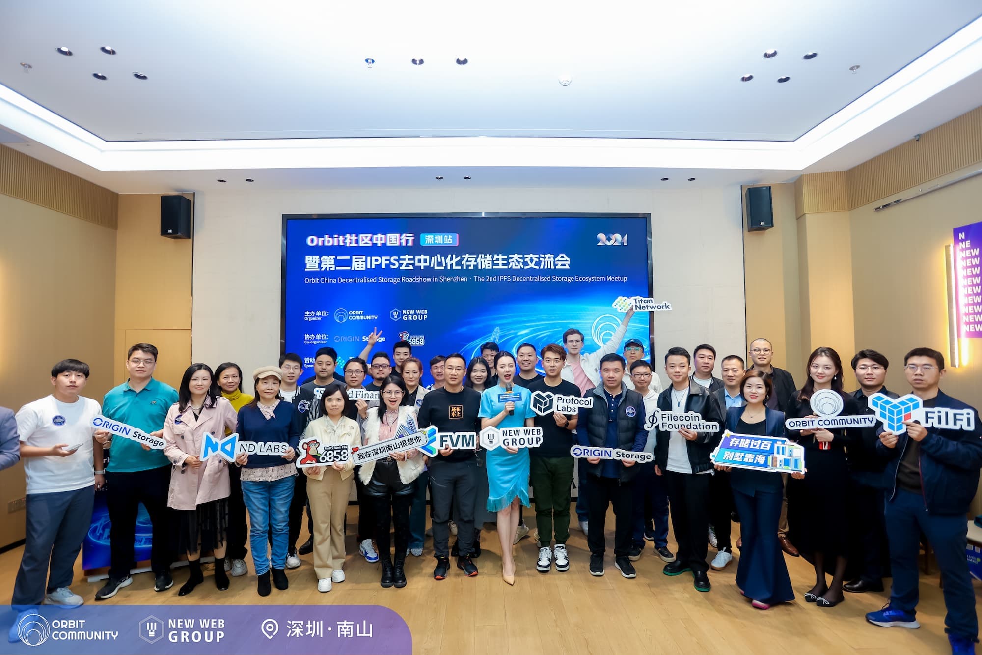 A large group of attendees posing for a group photo at the Orbit China Decentralized Storage Roadshow in Shenzhen. They are holding various signs with logos and names of different organizations like Filecoin Foundation, Protocol Labs, ND Labs, and Origin Storage. The background displays a large banner with the event name and sponsors, in both Chinese and English. The attendees are smiling and standing together in a well-lit conference room.